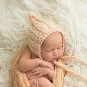 May include: A newborn baby wearing a knitted beige bonnet with long ties, wrapped in a matching blanket, and lying on a fluffy white rug. The baby is sleeping peacefully, curled up in a comfortable position.