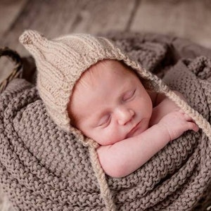 Peut inclure: Un nouveau-né portant un bonnet en tricot beige dort paisiblement dans une couverture en tricot marron. Le bébé est emmailloté dans la couverture et a une expression paisible sur son visage.