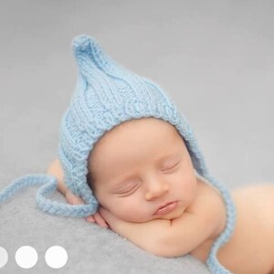 May include: A newborn baby wearing a light blue knitted hat with a long tassel. The baby is sleeping on a soft, gray blanket.