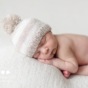 Pode incluir: Um bebê recém-nascido usando um chapéu de tricô branco e marrom com um pompom dorme em um cobertor branco.