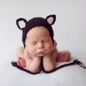 Puede incluir: Un bebé recién nacido con un gorro de gato de punto negro y blanco con orejas. El bebé está durmiendo con las manos debajo de la barbilla.