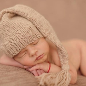 May include: A newborn baby wearing a beige knitted hat with a long tassel sleeping on a brown background.