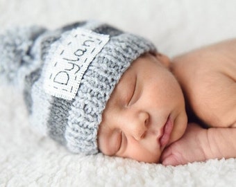 Pode incluir: Um gorro de bebé de malha listrada em cinza e branco com uma etiqueta branca que diz "Dylan" em letras pretas.