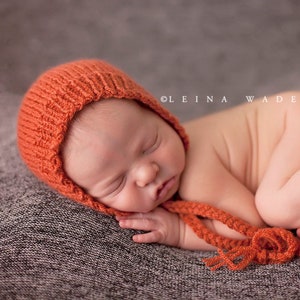 Può includere: Un neonato che indossa un berretto di lana arancione con un lungo fiocco. Il bambino sta dormendo su una superficie grigia testurizzata.