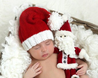 May include: A newborn baby wearing a red and white knitted Santa hat sleeps in a wicker basket filled with white faux fur. A small Santa doll is lying next to the baby.