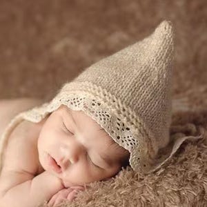 May include: A sleeping newborn baby wearing a beige knitted bonnet with a lace trim, lying on a brown, textured surface. The baby's hands are near its face. The image includes the text "Little New York photography".