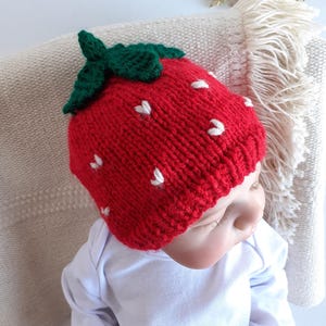 May include: A red knitted strawberry hat with green leaves and white seed details. The hat is worn by a baby wearing a white onesie. The hat is a novelty item and is likely handmade.