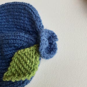 May include: A handmade blue knit hat with a green leaf accent. The hat is made of soft yarn and features a textured design. The leaf is a vibrant green color, adding a pop of color to the hat. This hat is perfect for babies and toddlers.