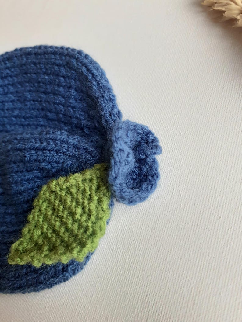 May include: A handmade blue knit hat with a green leaf accent. The hat is made of soft yarn and features a textured design. The leaf is a vibrant green color, adding a pop of color to the hat. This hat is perfect for babies and toddlers.