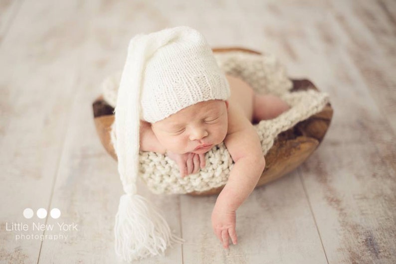 May include: A newborn baby wearing a white knitted hat with a long tassel, sleeping in a wooden bowl lined with white crocheted fabric.