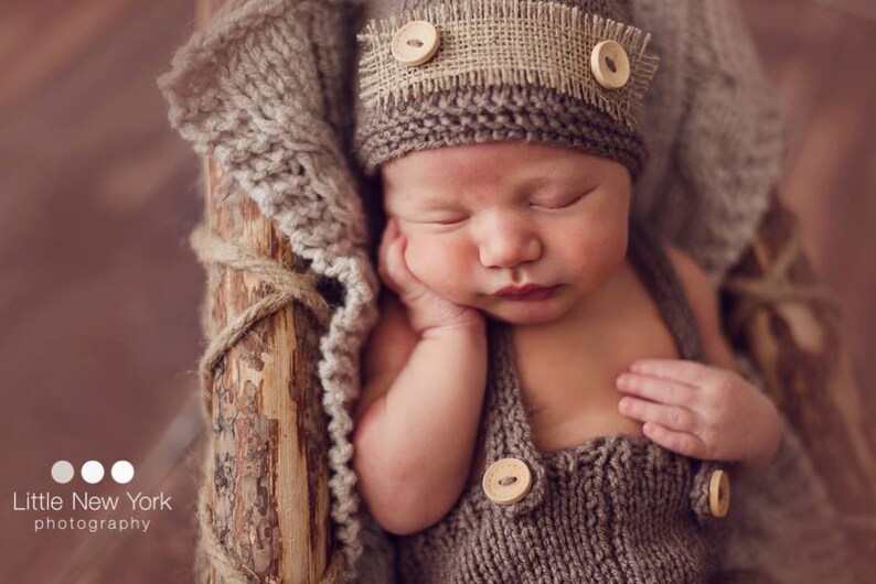 K&ouml;nnte beinhalten: Ein schlafendes Baby tr&auml;gt eine braune Strickm&uuml;tze mit einem Leinenband und Kn&ouml;pfen. Das Baby tr&auml;gt ein braunes Strickoutfit mit Kn&ouml;pfen. Das Baby liegt auf einer braunen Strickdecke.