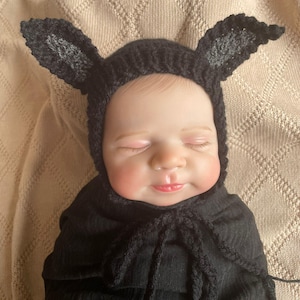 May include: A baby wearing a black knitted bunny hat with long ears and a matching black wrap. The baby is lying on a beige knitted blanket. The baby's eyes are closed, and the lips are slightly parted.
