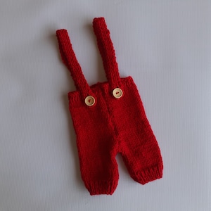 May include: A bright red knitted garment featuring two shoulder straps and two light-colored wooden buttons attached to the front waistband. The piece is constructed with a textured knit pattern, including ribbed cuffs at the bottom of the legs.
