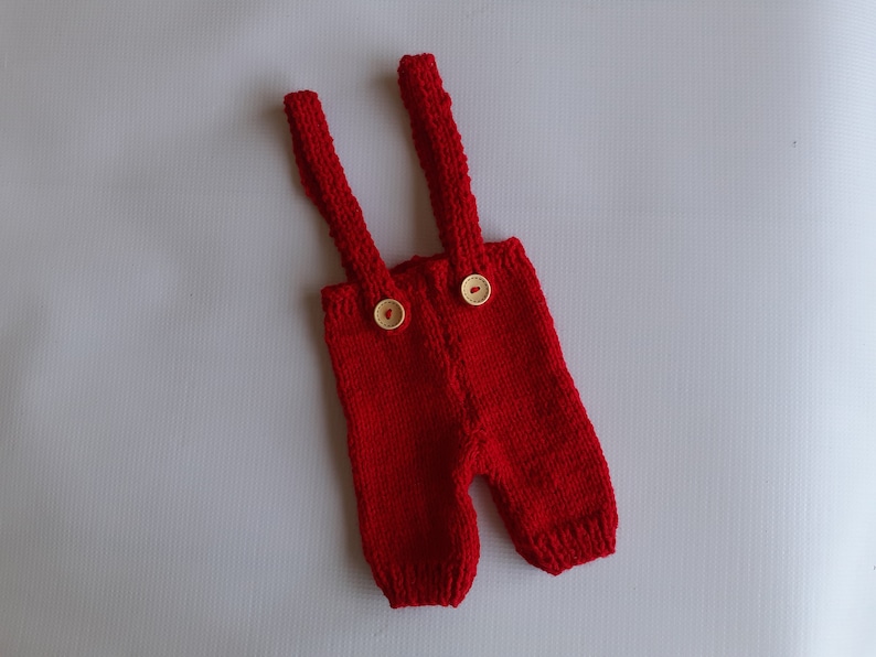 May include: A bright red knitted garment featuring two shoulder straps and two light-colored wooden buttons attached to the front waistband. The piece is constructed with a textured knit pattern, including ribbed cuffs at the bottom of the legs.