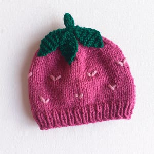 May include: A knitted strawberry hat in shades of pink and green. The hat features a ribbed brim, a pink body with small pink seed details, and a green leaf and stem design on top. This handmade hat is perfect for babies and children.