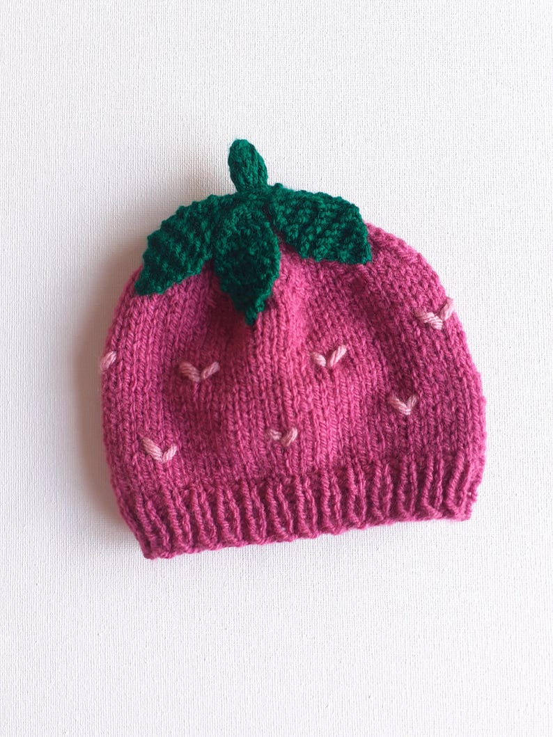May include: A knitted strawberry hat in shades of pink and green. The hat features a ribbed brim, a pink body with small pink seed details, and a green leaf and stem design on top. This handmade hat is perfect for babies and children.