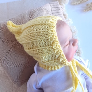 May include: A knitted yellow baby bonnet with ear flaps and tie strings. The bonnet is made of a textured yarn and is shown on a baby. The background includes a beige blanket and a white surface.