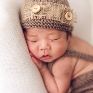 K&ouml;nnte beinhalten: Ein schlafendes Baby tr&auml;gt eine braune Strickm&uuml;tze mit einem Band aus Sackleinen und Holzkn&ouml;pfen. Das Baby ist in eine braune Strickdecke gewickelt.