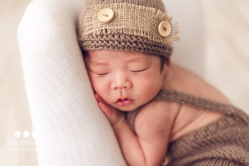 K&ouml;nnte beinhalten: Ein schlafendes Baby tr&auml;gt eine braune Strickm&uuml;tze mit einem Band aus Sackleinen und Holzkn&ouml;pfen. Das Baby ist in eine braune Strickdecke gewickelt.