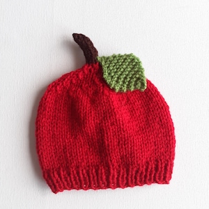 Pode incluir: Um gorro de malha vermelho brilhante em forma de maçã, com uma borda canelada na parte inferior, um caule castanho curto que sobressai do centro superior e uma única folha verde texturizada presa junto ao caule no lado superior direito.
