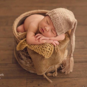 May include: A newborn baby sleeping in a brown wicker basket, wearing a beige knitted hat with a long tassel. The baby is wrapped in a brown and yellow knitted blanket.