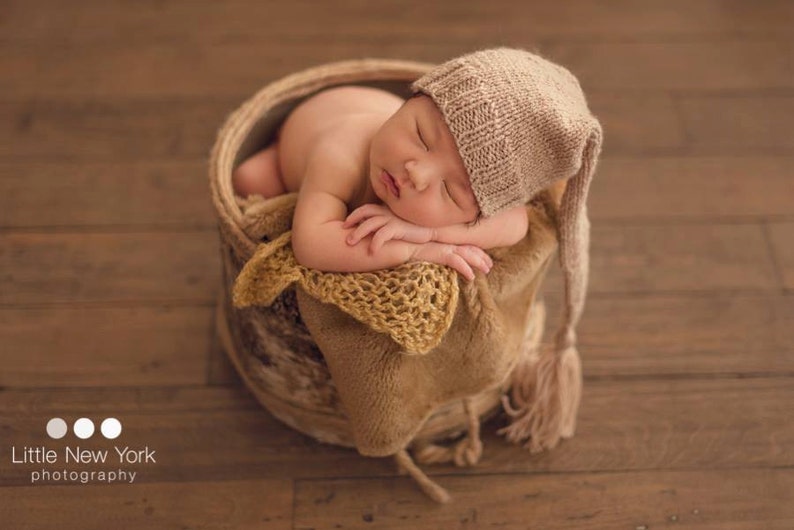May include: A newborn baby sleeping in a brown wicker basket, wearing a beige knitted hat with a long tassel. The baby is wrapped in a brown and yellow knitted blanket.