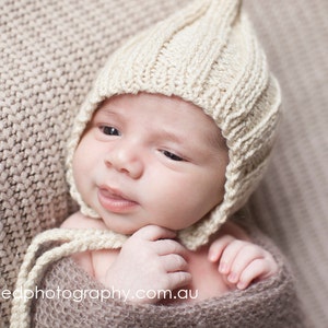 Peut inclure: Un nouveau-né portant un bonnet en tricot de couleur crème avec un sommet pointu. Le bébé est allongé sur une couverture en tricot marron.