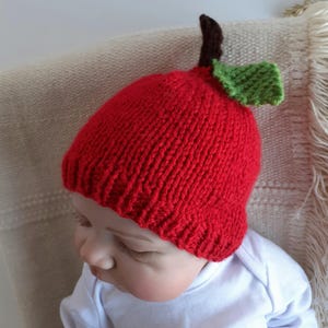 May include: A red knitted baby hat shaped like an apple, complete with a brown stem and green leaf. The hat is worn by a baby wearing a white onesie. The hat is a fun accessory for babies.