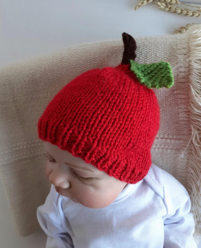 May include: A red knitted baby hat shaped like an apple, complete with a brown stem and green leaf. The hat is worn by a baby wearing a white onesie. The hat is a fun accessory for babies.