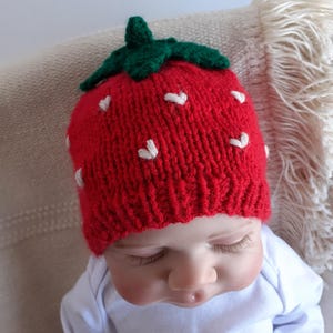 May include: A red knitted strawberry hat with white heart-shaped accents and a green stem. The hat is worn by a baby wearing a white onesie.
