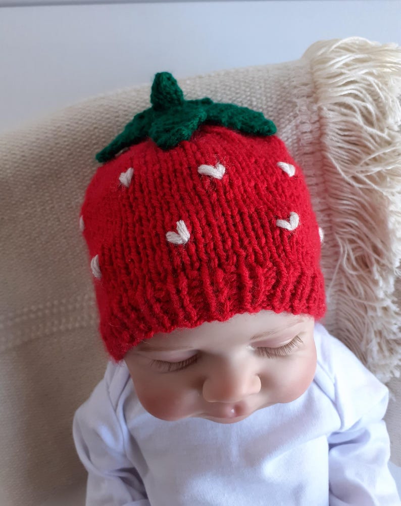 May include: A red knitted strawberry hat with white heart-shaped accents and a green stem. The hat is worn by a baby wearing a white onesie.