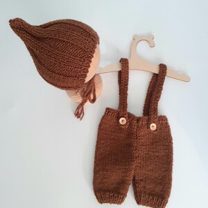 May include: A brown knitted baby outfit, including a bonnet and overalls with wooden buttons, displayed on a wooden hanger. The outfit is a warm, earthy brown color, perfect for newborn photography or a baby shower gift.