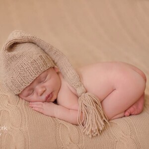 May include: A newborn baby wearing a beige knitted hat with a long tassel, sleeping on a beige knitted blanket.