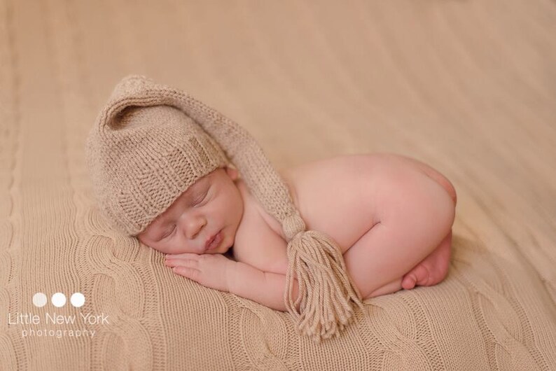 May include: A newborn baby wearing a beige knitted hat with a long tassel, sleeping on a beige knitted blanket.