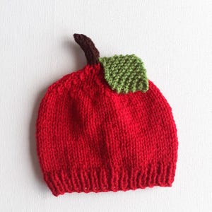 May include: A knitted red baby hat shaped like an apple, complete with a brown stem and a green leaf. The hat is made of soft yarn and is designed to resemble a realistic apple. The hat is a fun and whimsical accessory.