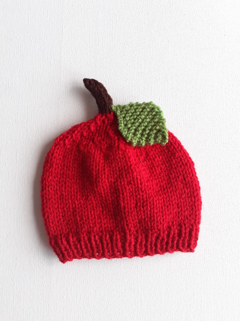 May include: A knitted red baby hat shaped like an apple, complete with a brown stem and a green leaf. The hat is made of soft yarn and is designed to resemble a realistic apple. The hat is a fun and whimsical accessory.