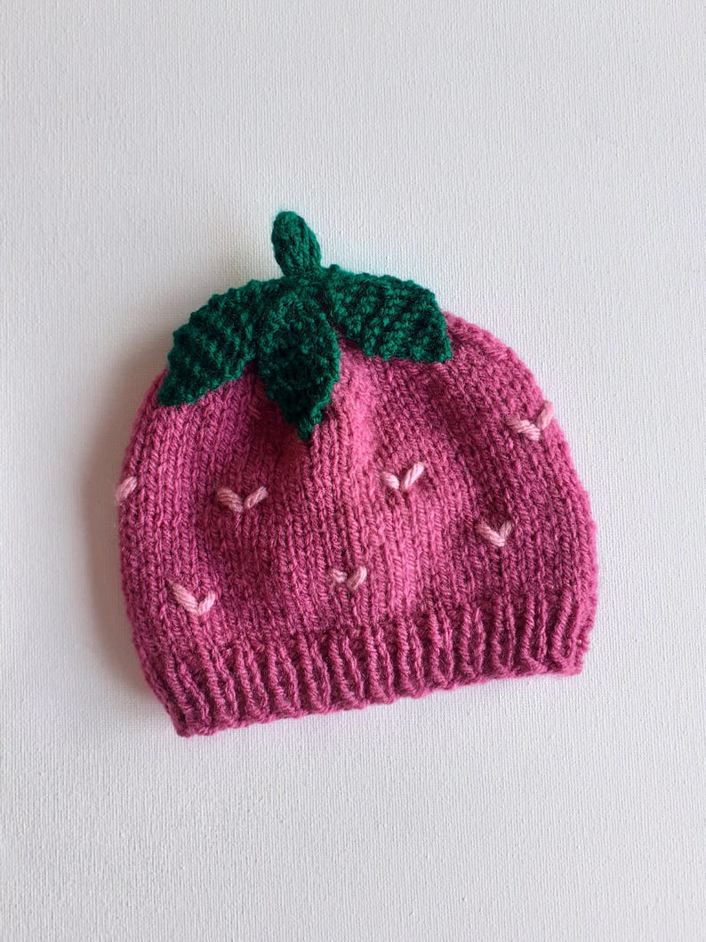 May include: A knitted hat in the shape of a strawberry. The hat is primarily pink with green leaves and a stem at the top. Small pink stitches are scattered across the hat, resembling strawberry seeds.
