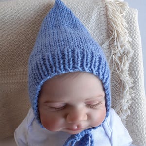 May include: A pale blue knitted baby bonnet with a pointed crown and ties. The bonnet is made of soft yarn. The baby bonnet is designed to keep a baby's head warm. The bonnet is shown on a baby wearing a white onesie.