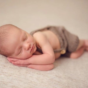 K&ouml;nnte beinhalten: Ein neugeborenes Baby schl&auml;ft auf einer beigefarbenen Decke und tr&auml;gt ein braunes Strickoutfit mit einem Knopf.