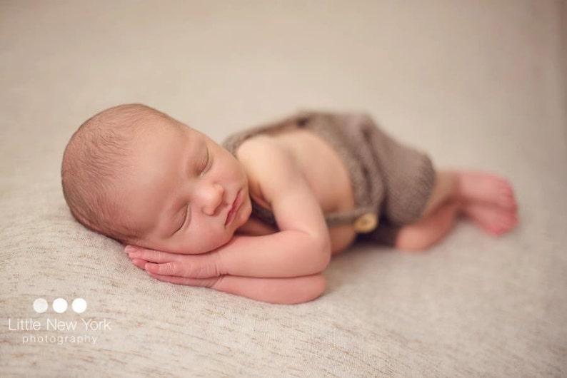 K&ouml;nnte beinhalten: Ein neugeborenes Baby schl&auml;ft auf einer beigefarbenen Decke und tr&auml;gt ein braunes Strickoutfit mit einem Knopf.