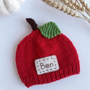 May include: A knitted red baby hat shaped like an apple, with a green leaf and brown stem. A white patch with the name "Ben" is sewn on the front. The hat is made of a soft, textured yarn.