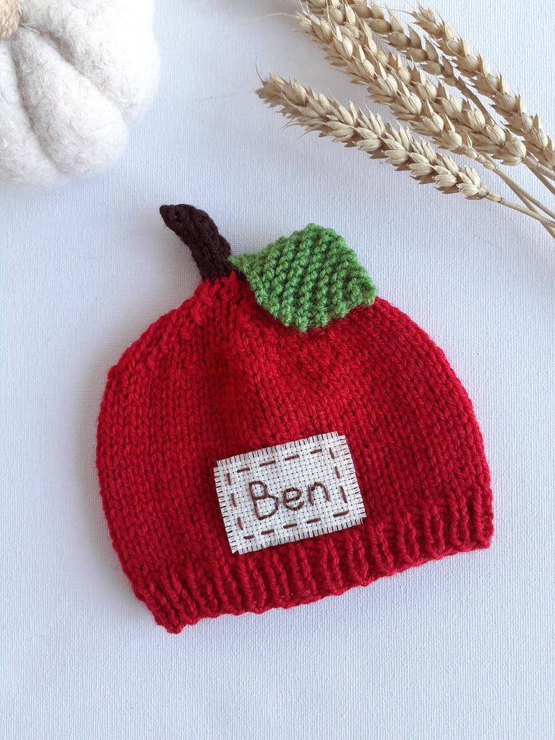 May include: A knitted red baby hat shaped like an apple, with a green leaf and brown stem. A white patch with the name "Ben" is sewn on the front. The hat is made of a soft, textured yarn.