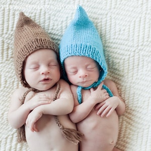 Op de afbeelding: Twee slapende pasgeborenen met gebreide puntmutsen, één bruin en één blauw, liggen naast elkaar op een witte, getextureerde deken. De mutsen hebben strikjes. De baby's hebben de armen gekruist.