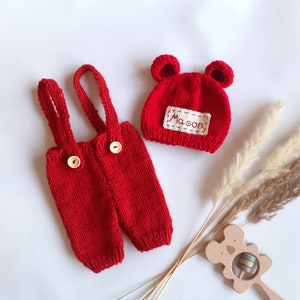 May include: A bright red knitted overall set with two shoulder straps and two light-colored wooden buttons on the front. The set includes a matching red knitted hat with two small rounded ears and a white rectangular patch featuring the name Mason.