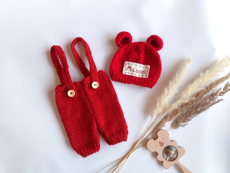 May include: A bright red knitted overall set with two shoulder straps and two light-colored wooden buttons on the front. The set includes a matching red knitted hat with two small rounded ears and a white rectangular patch featuring the name Mason.
