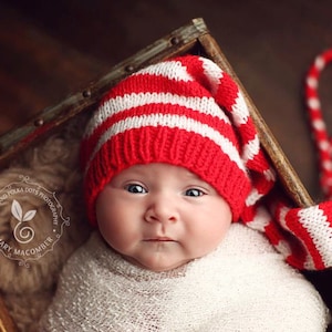 May include: A baby wearing a red and white striped knitted hat with a long tail. The hat is pulled down over the baby's eyes. The baby is lying in a wooden box.
