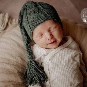 May include: A baby wearing a dark green knitted hat with a long tassel, and a white onesie. The baby is lying down, eyes closed, and smiling slightly. The background is a soft, neutral colour.