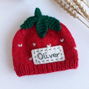 May include: A red knit baby hat shaped like a strawberry, with green leaves and white heart accents. The name "Oliver" is embroidered on a white patch. The hat is designed for infants and toddlers.