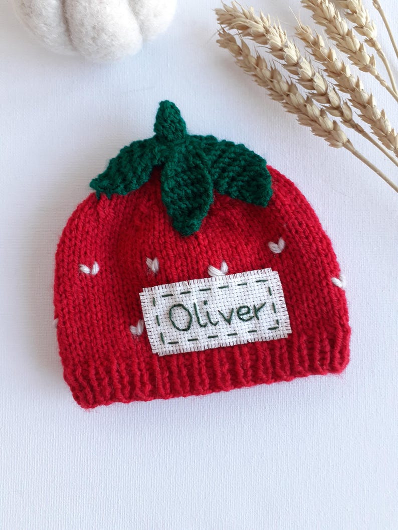 May include: A red knit baby hat shaped like a strawberry, with green leaves and white heart accents. The name "Oliver" is embroidered on a white patch. The hat is designed for infants and toddlers.