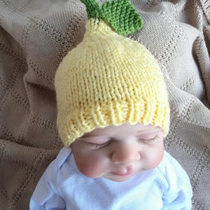 May include: A knitted baby hat in the shape of a lemon, with a green leaf detail. The hat is yellow and sits on a baby wearing a white onesie. The baby is lying on a beige blanket.